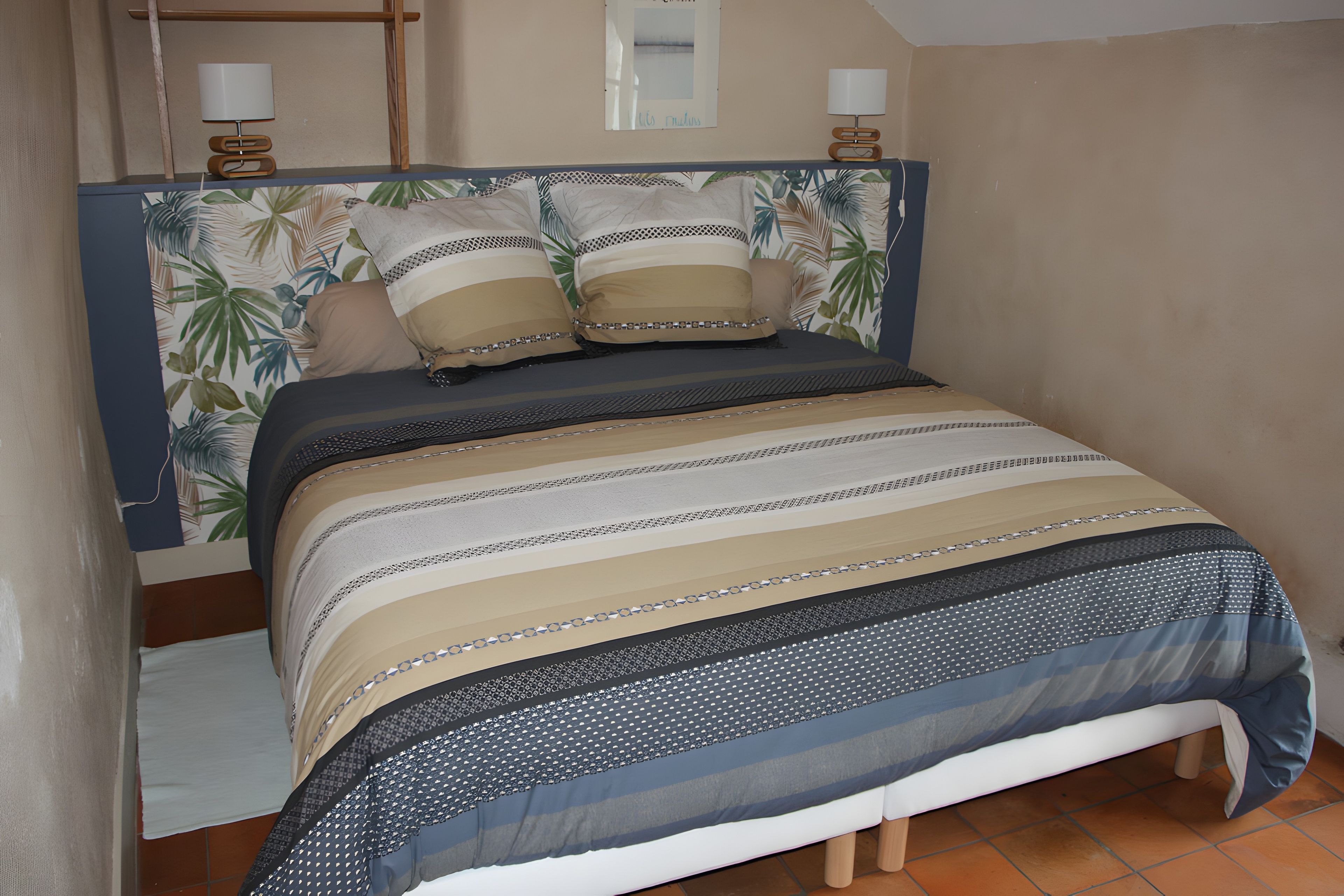 Uma cama com cabeceira estampada, duas luminárias de apoio e uma parede com um quadro emoldurado.