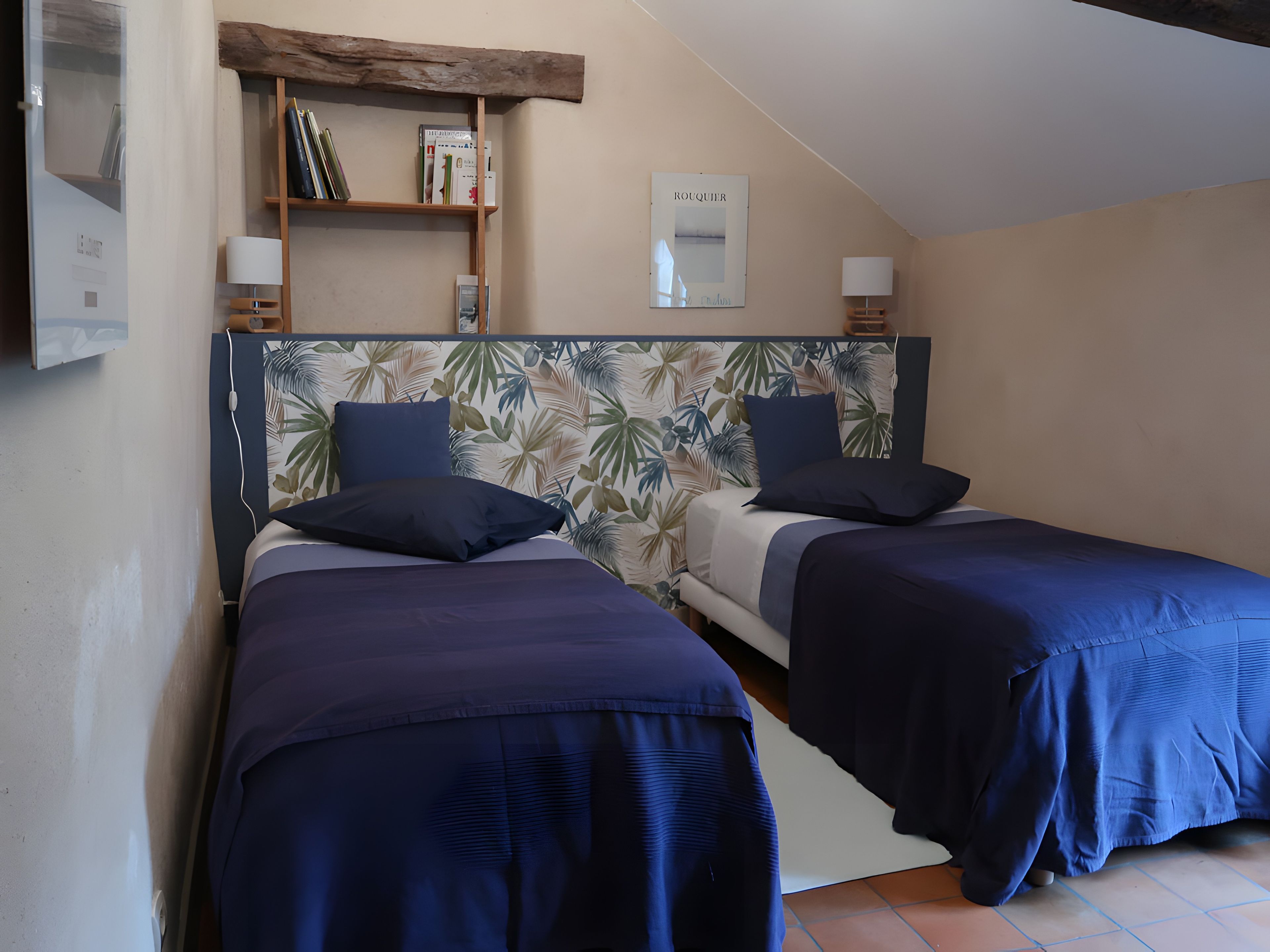 Quarto com duas camas, uma estante de madeira com livros, um espelho e um candeeiro fixado na parede.