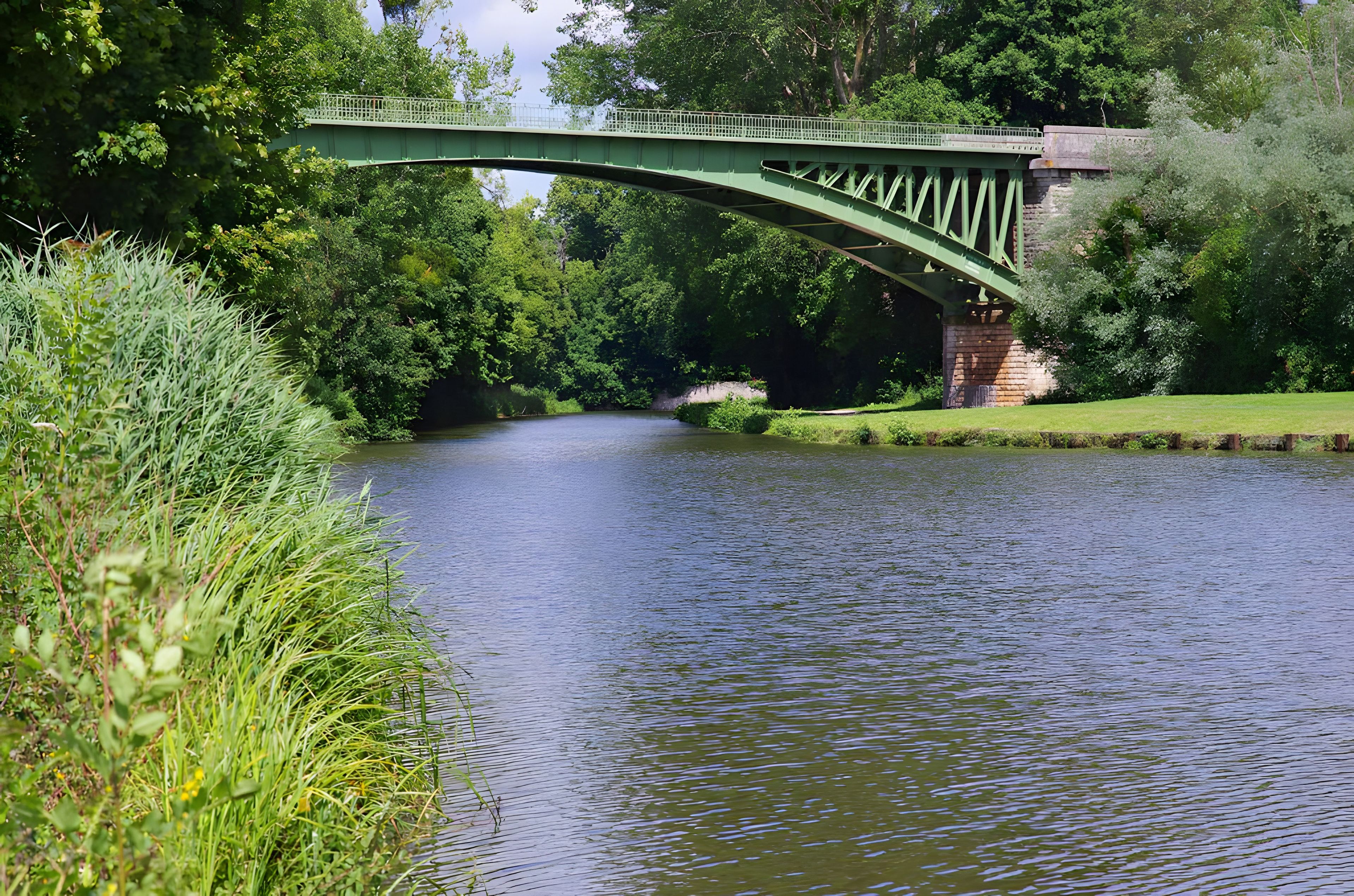 A green metal bridge over a calm river, surrounded by lush greenery and trees.