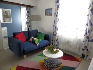 Uma sala de estar com um sofá azul, almofadas coloridas, uma mesa branca com uma planta e um tapete geométrico.