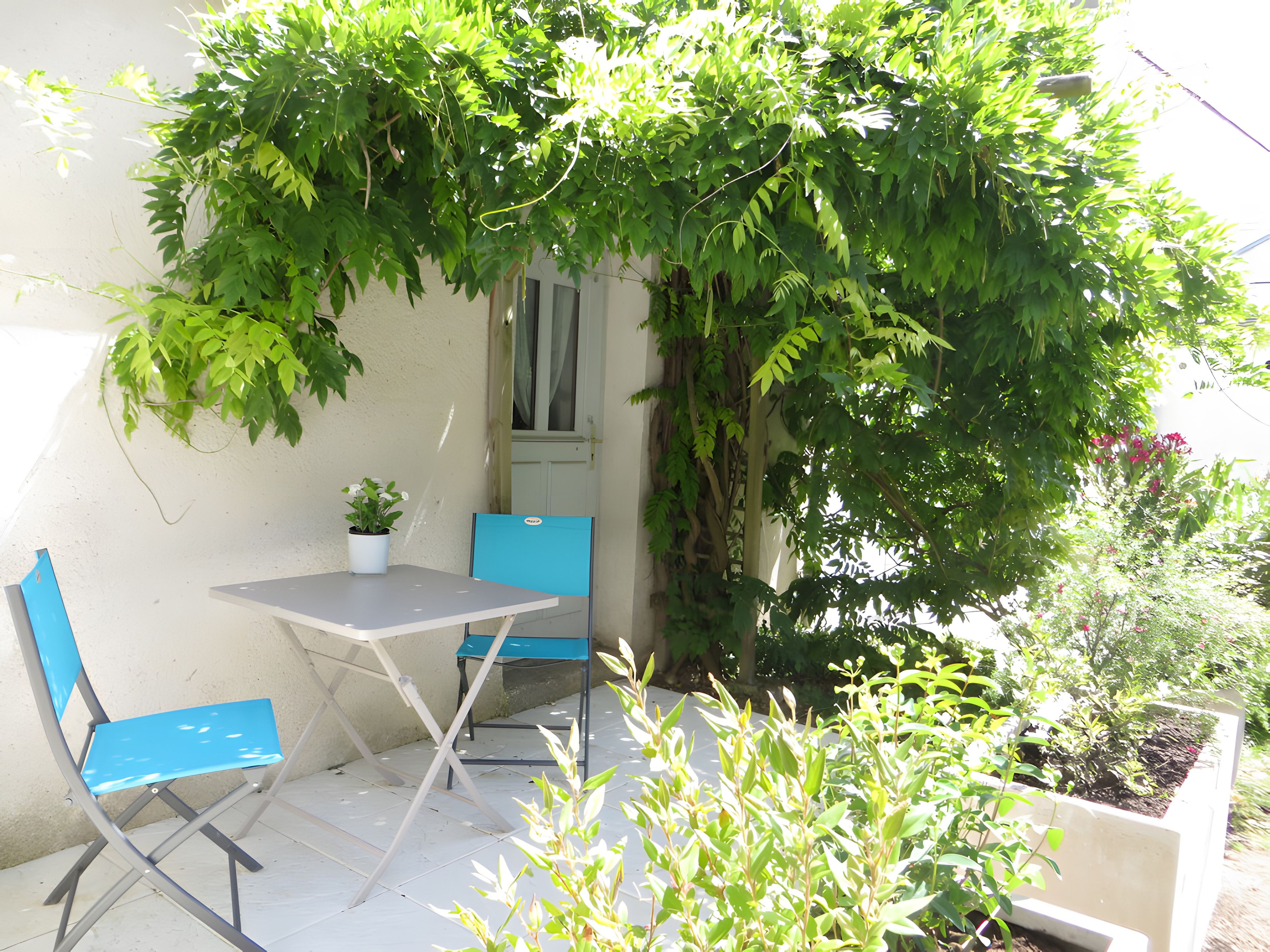 Área externa com mesa, duas cadeiras e vegetação, cercada por um muro branco.