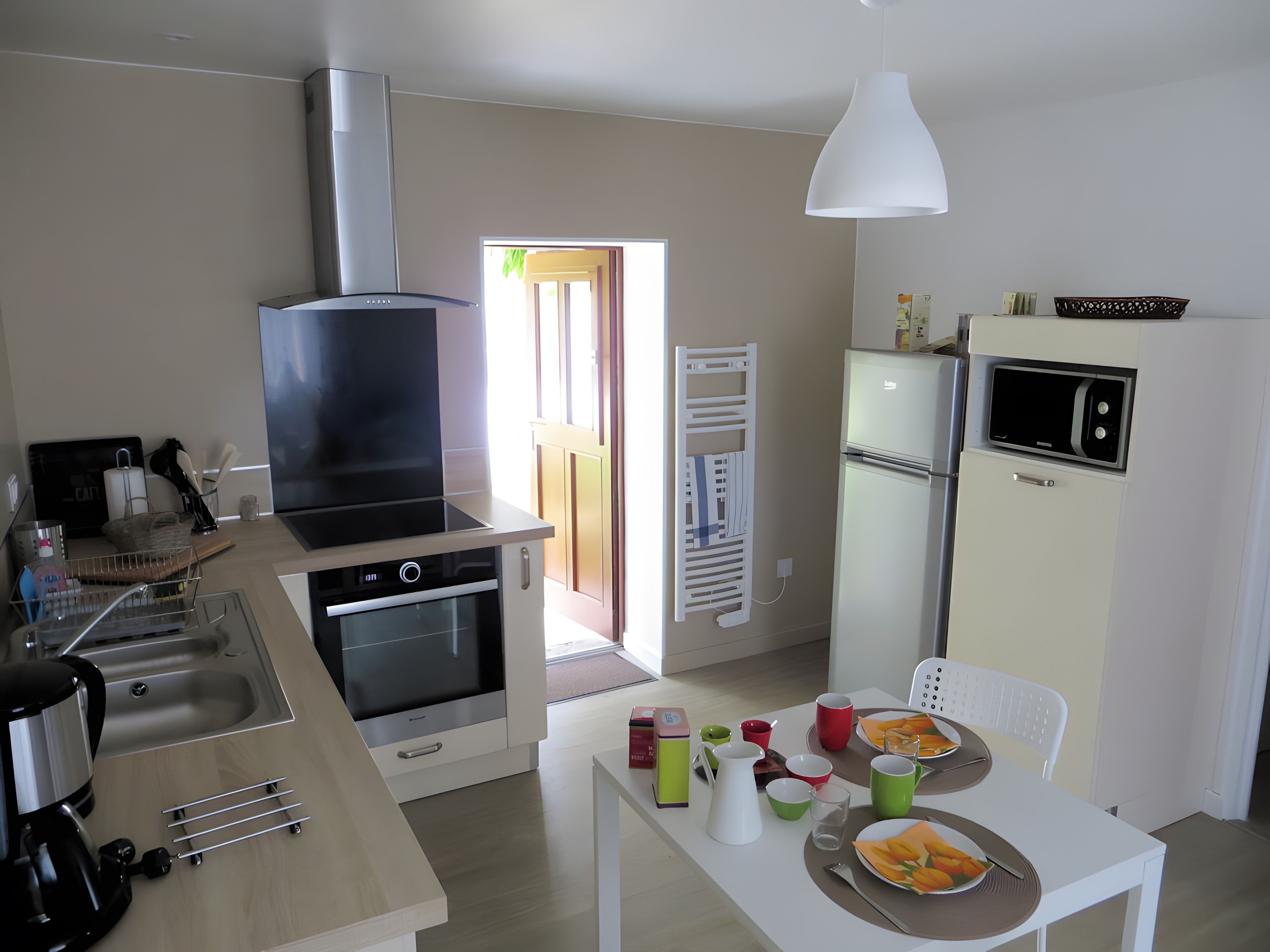Een moderne keuken met een ingebouwde oven, magnetron en een groot fornuis. Er is een eethoek met een tafel voor twee personen, en een koelkast met een magnetron erboven.