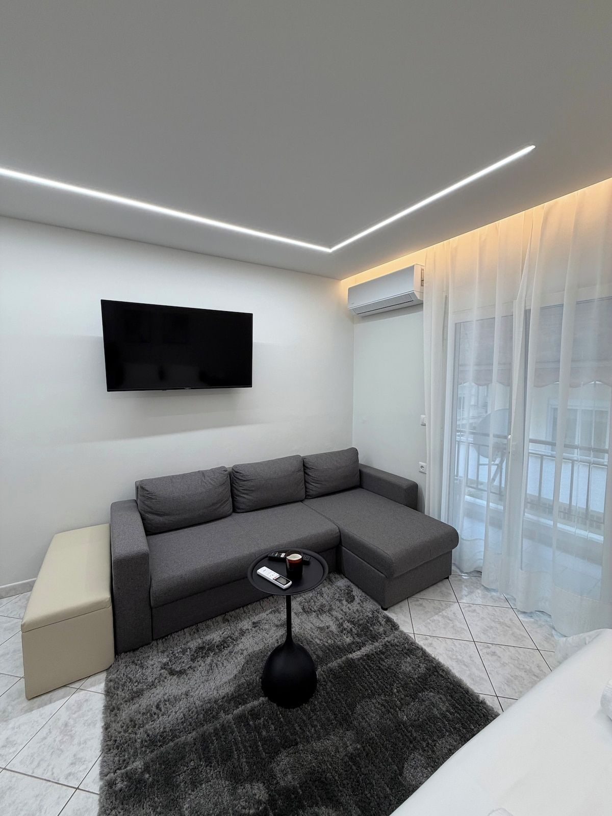 Un salon moderne avec un canapé d’angle, un téléviseur à écran plat fixé au mur, une petite table basse ronde et un lampadaire noir.
