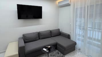 Un salon moderne avec un canapé d’angle, un téléviseur à écran plat fixé au mur, une petite table basse ronde et un lampadaire noir.