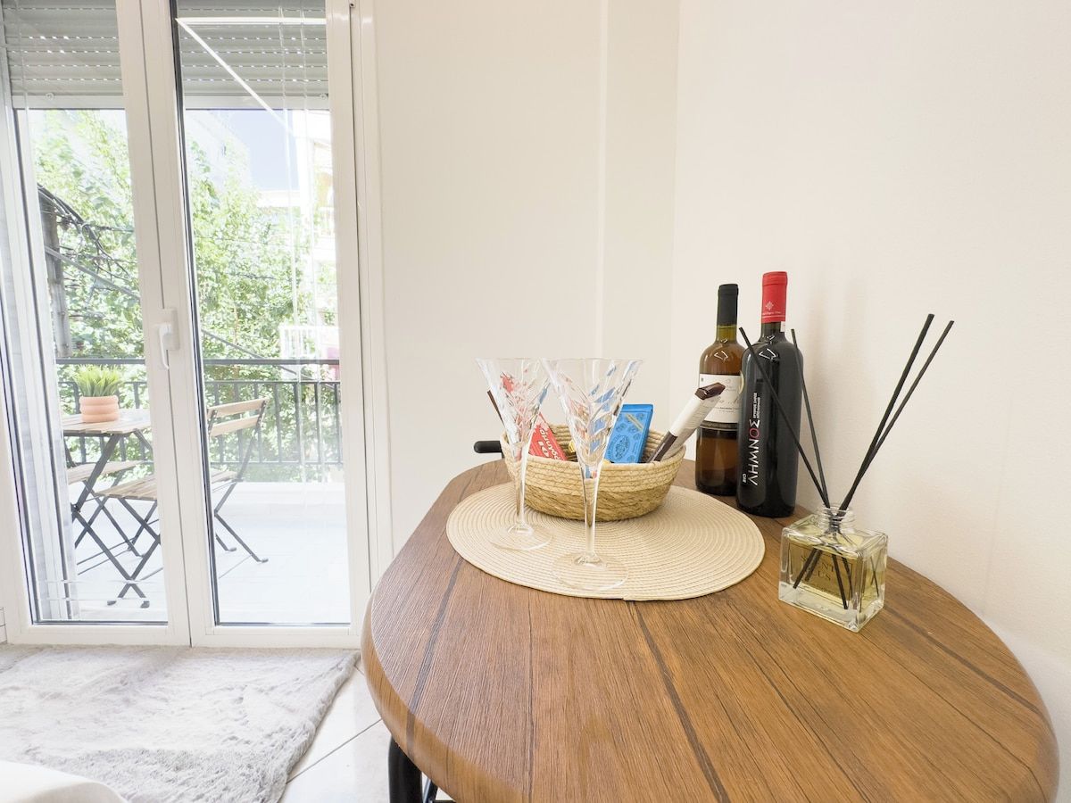 A wooden table with a woven placemat, a glass vase with reed sticks, and a basket containing books and a bottle of wine.