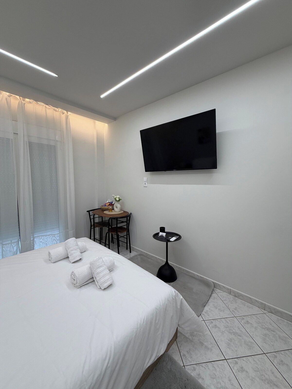 Ein Hotelzimmer mit einem Bett, einem an der Wand montierten Fernseher, einem kleinen Tisch mit Stuhl und einem fenster mit Vorhängen.