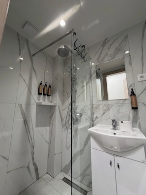 A modern bathroom with a glass-enclosed shower, marble walls, a white sink, and toiletries on a shelf. - Velórea Exclusive Residences - Ándrea studio (Thessaloniki)