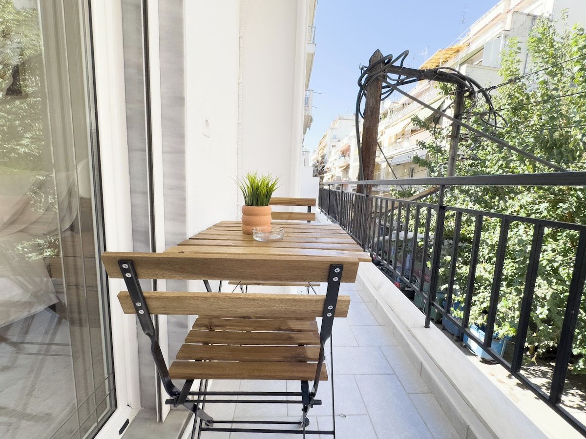 A balcony with a wooden table and benches, a potted plant, and a view of buildings and trees.