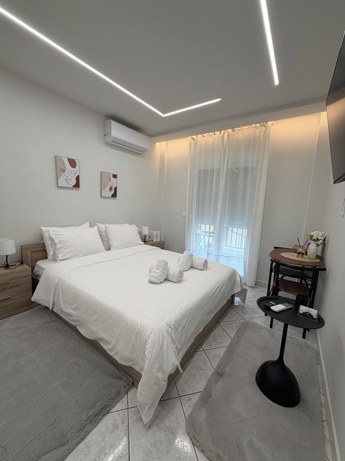 A modern bedroom with a large bed, bedside tables, a flat-screen TV, and a window with sheer curtains.