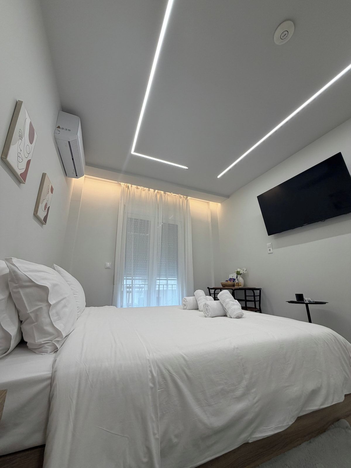 A modern hotel room with a bed, a flat-screen TV mounted on the wall, and a ceiling with linear lighting.