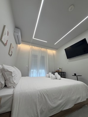 A modern hotel room with a bed, a flat-screen TV mounted on the wall, and a ceiling with linear lighting. - Velórea Exclusive Residences - Ándrea studio (Thessaloniki)