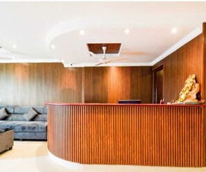 A reception area with a curved wooden reception desk, a ceiling fan, and a sofa in the background. - Classic Vaishnavi Residency (Hyderabad)