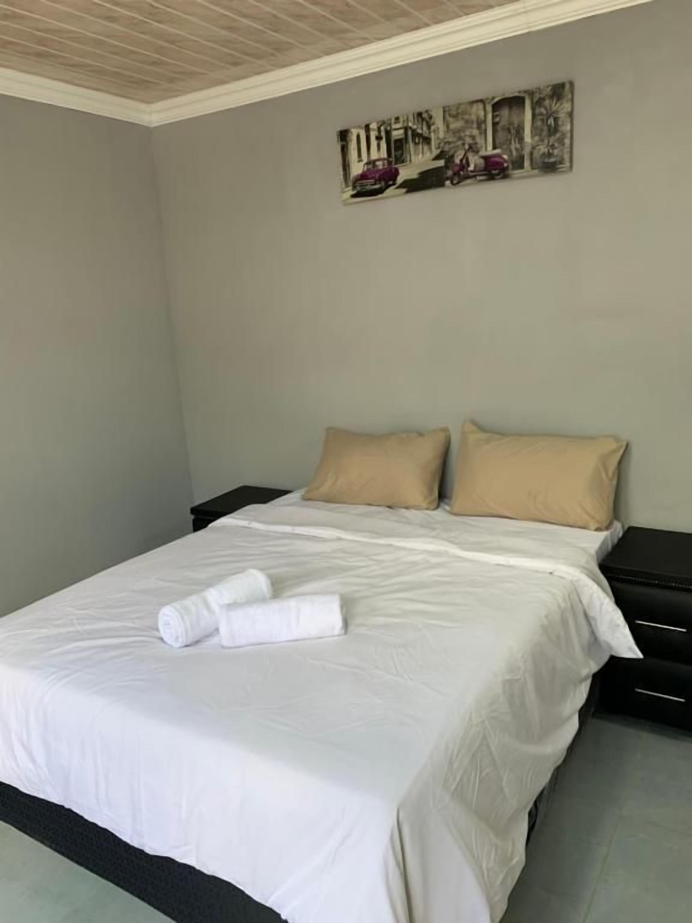 A neatly made bed with white linens and two beige pillows, a black nightstand, and a wall-mounted picture.