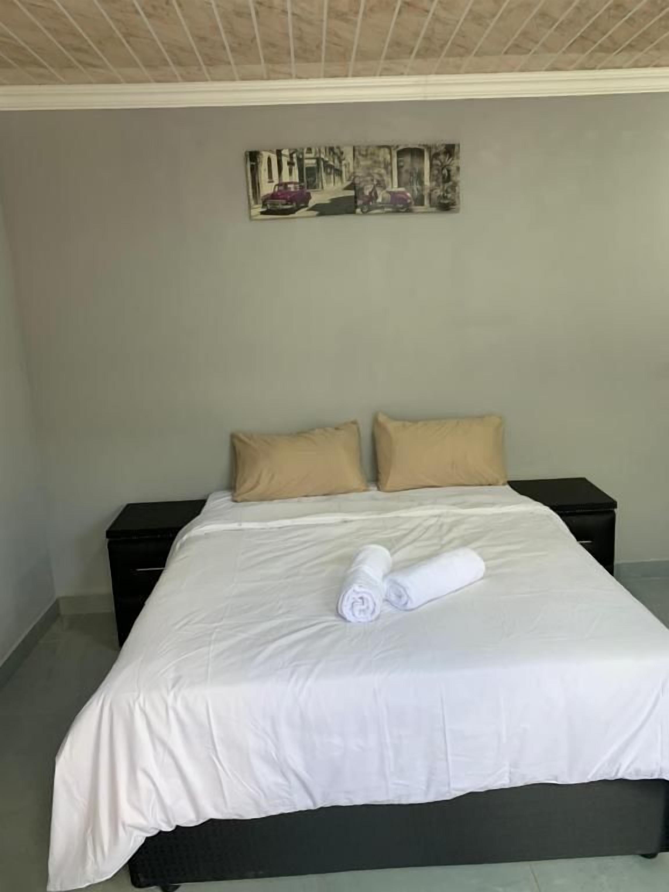 Una cama bien tendida con una toalla blanca enrollada, dos almohadas color beige y una mesita de noche negra al lado.