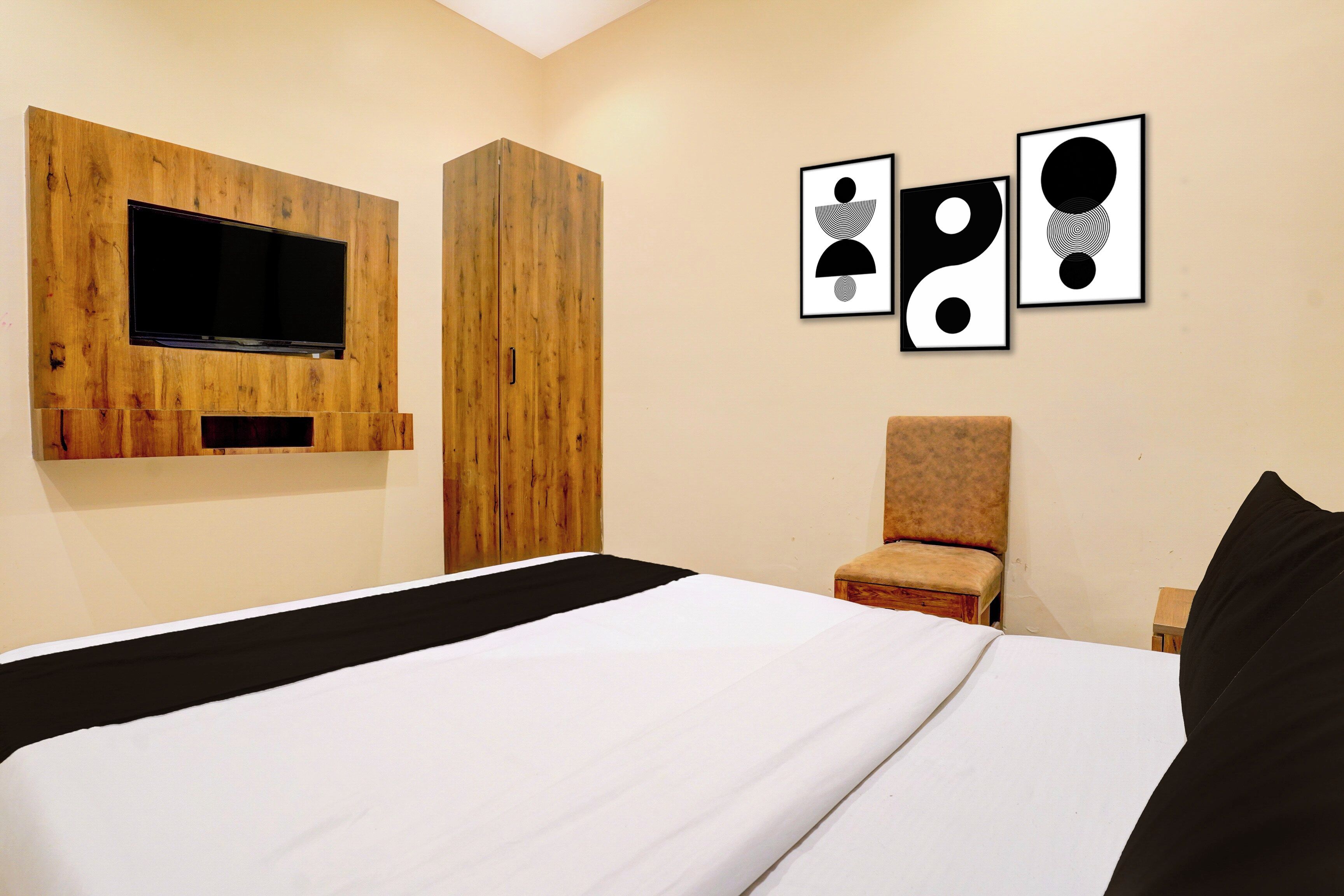 Habitación de hotel con cama, televisor montado en un panel de madera, una silla y tres cuadros en la pared.