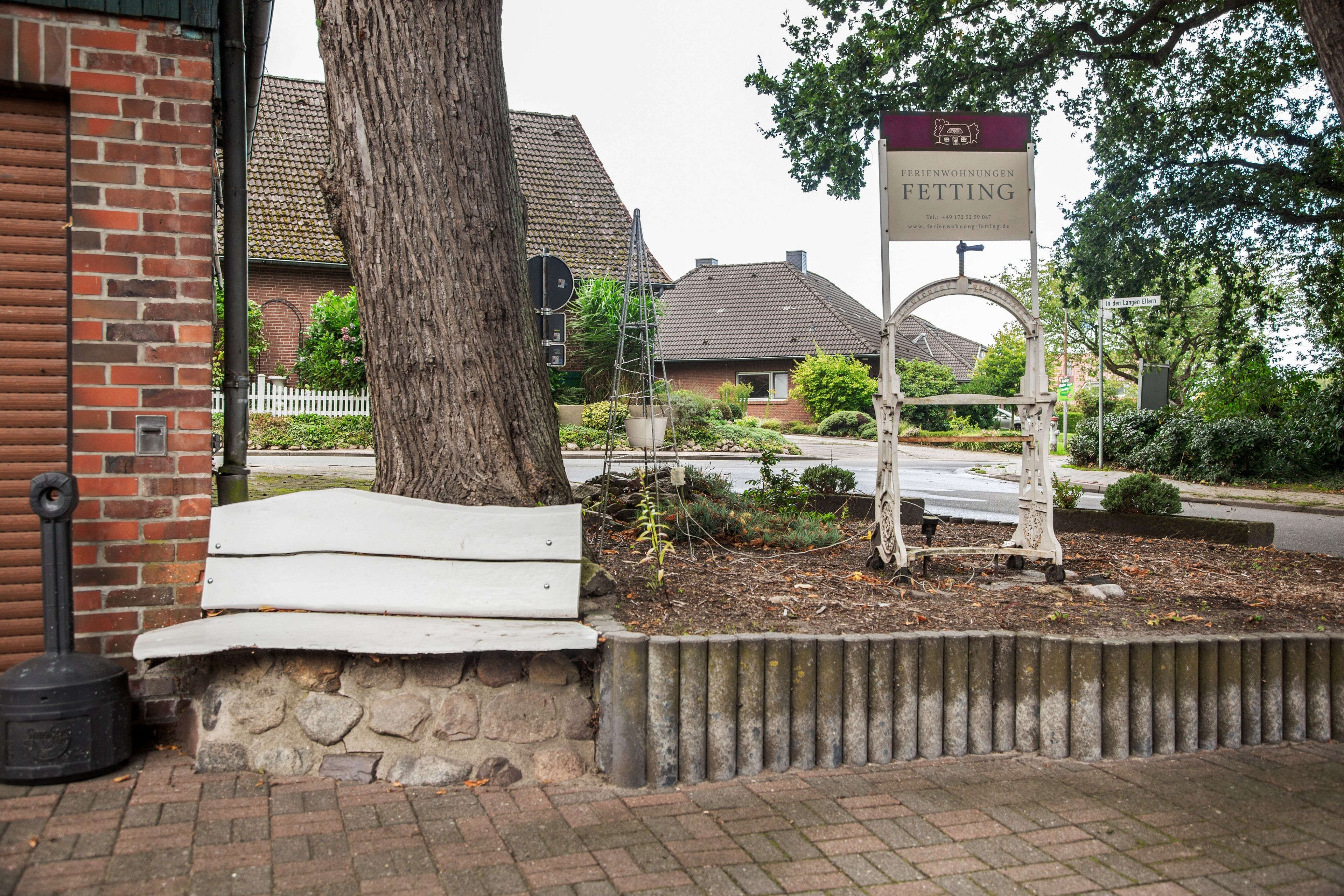 A brick building with a white bench, a tree, and a sign for 'Fetting' in front of a residential area.