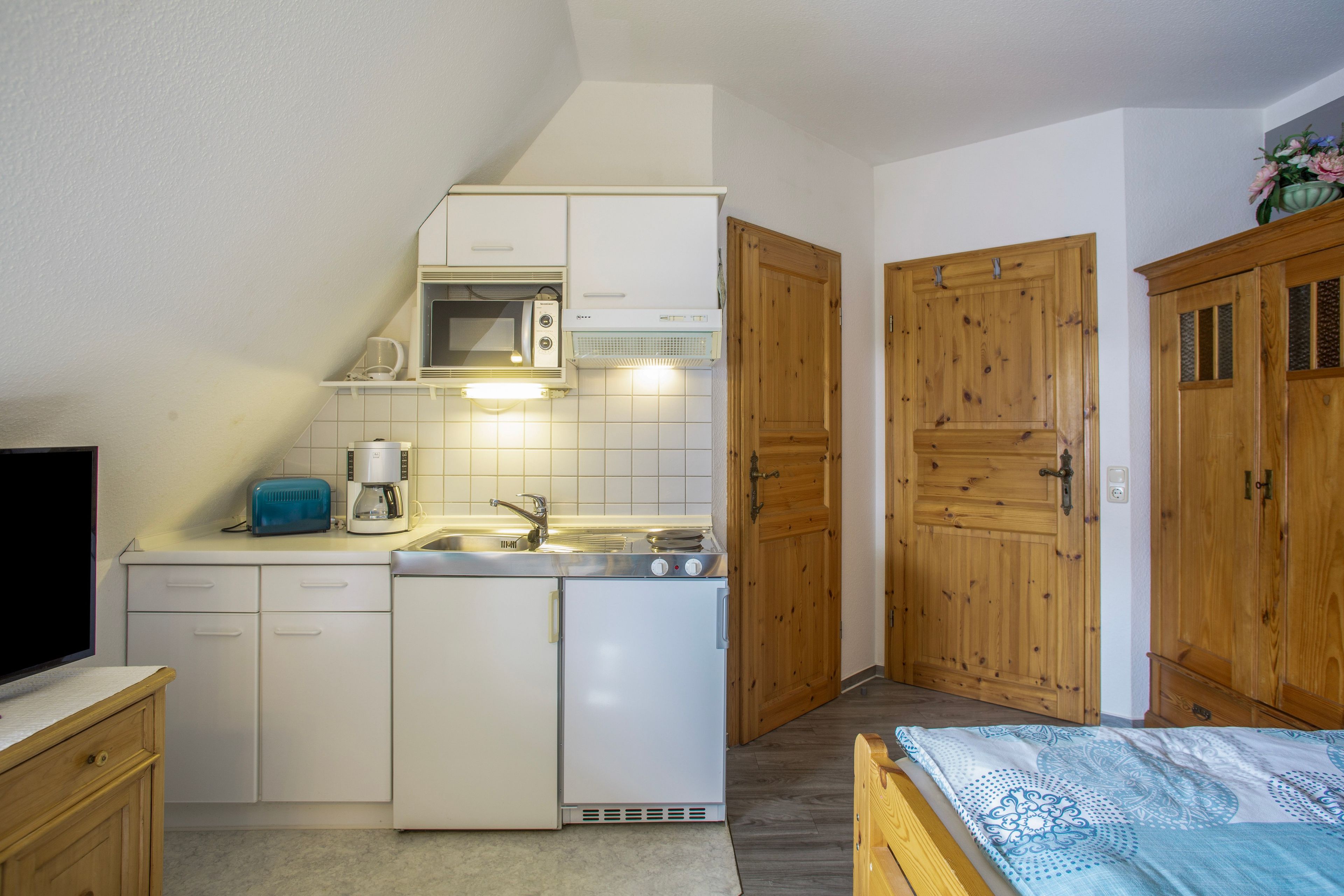 Eine kompakte Küche mit Mikrowelle, Toaster und Kaffeemaschine. Ein Spülbecken mit Wasserhahn ist vorhanden, sowie Holzschränke mit Glastüren. Im Hintergrund ist ein Bett mit einem gemusterten Überwurf zu sehen.