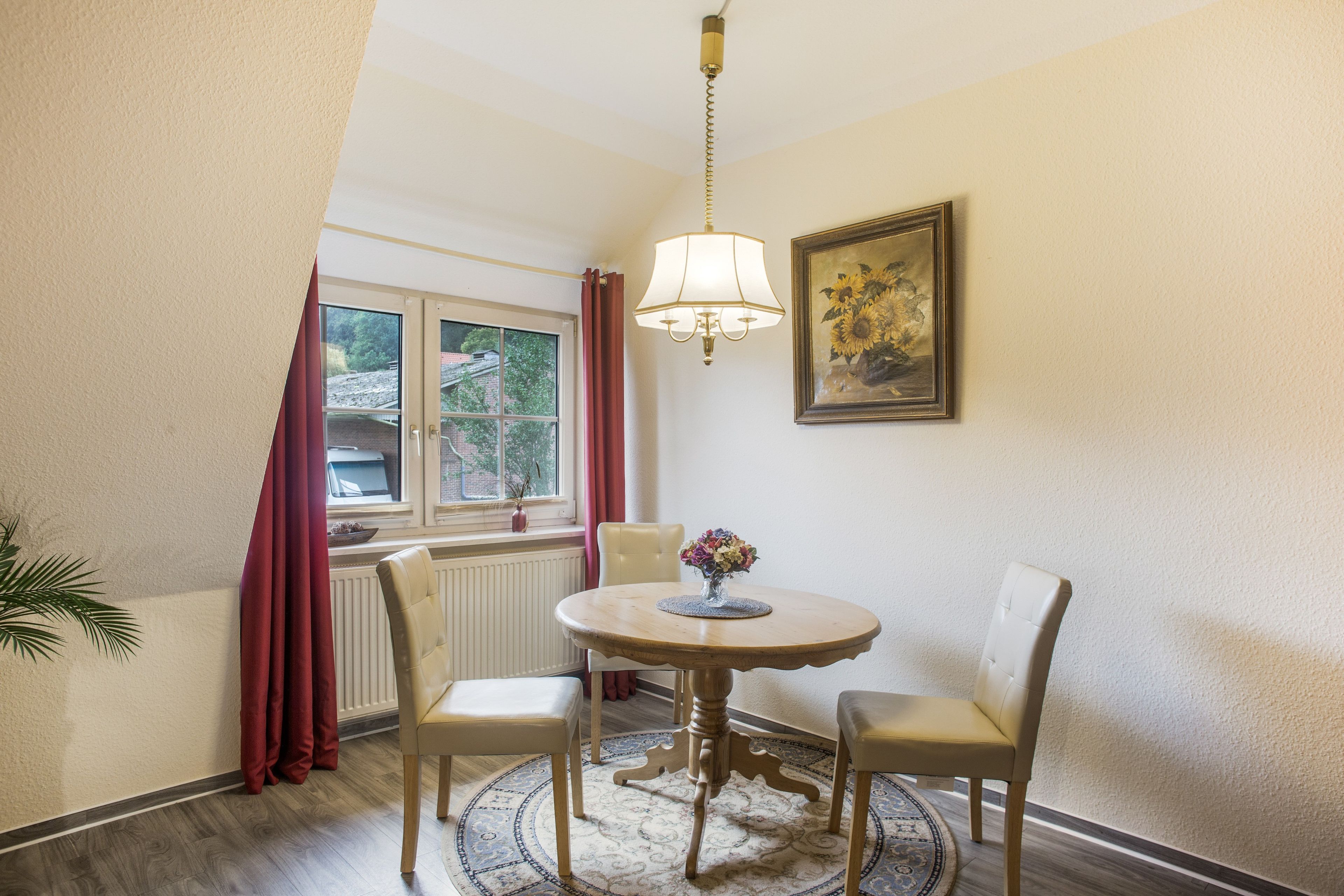 Ein Eckzimmer mit einem runden Tisch, zwei Stühlen, einem fenster mit Vorhängen, einem Heizkörper, einer Kristallleuchtreiter und einem gerahmten Gemälde an der Wand.