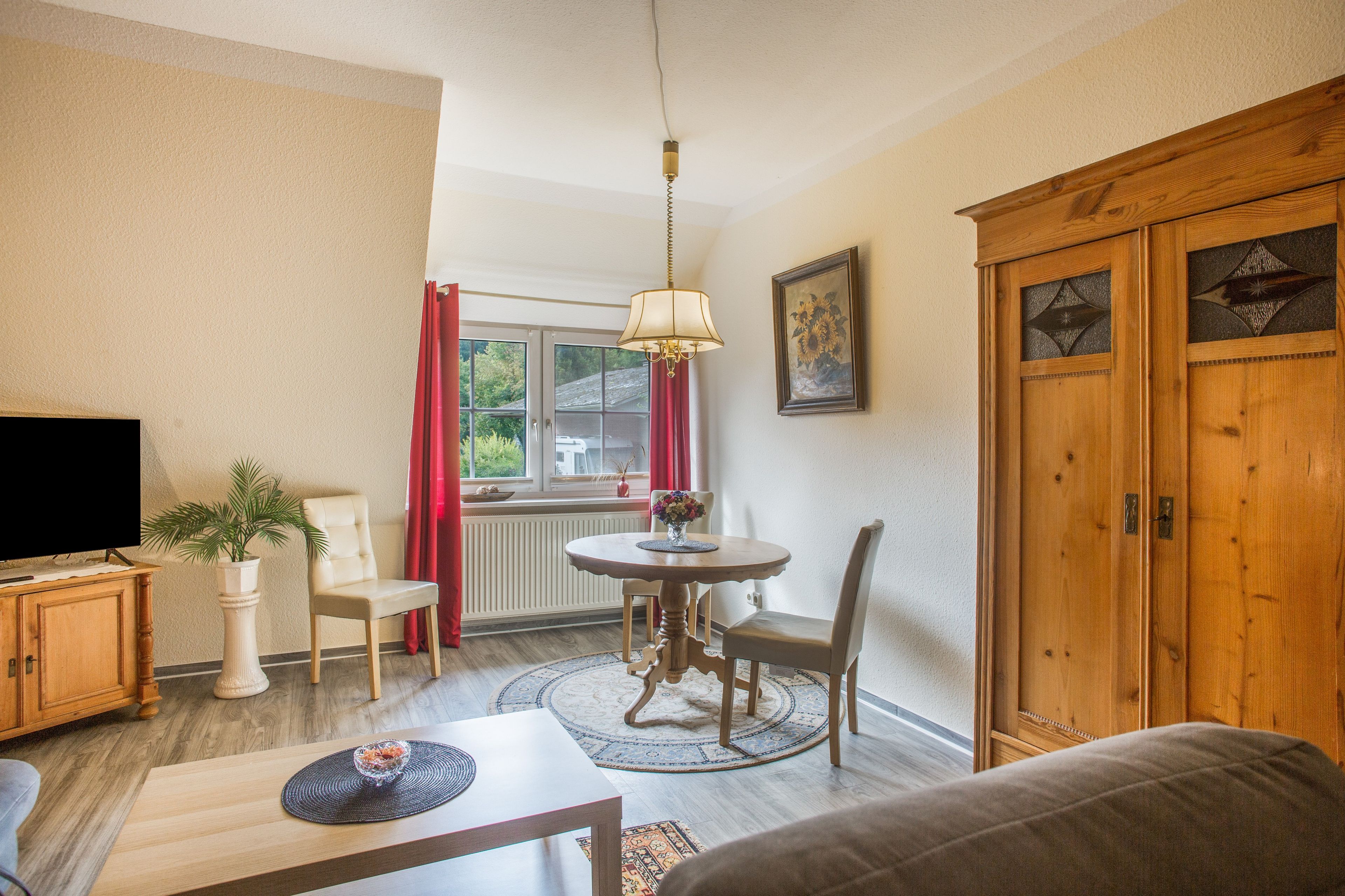 A living room with a television, a dining table with chairs, a sofa, a wooden cabinet, and a window with curtains.