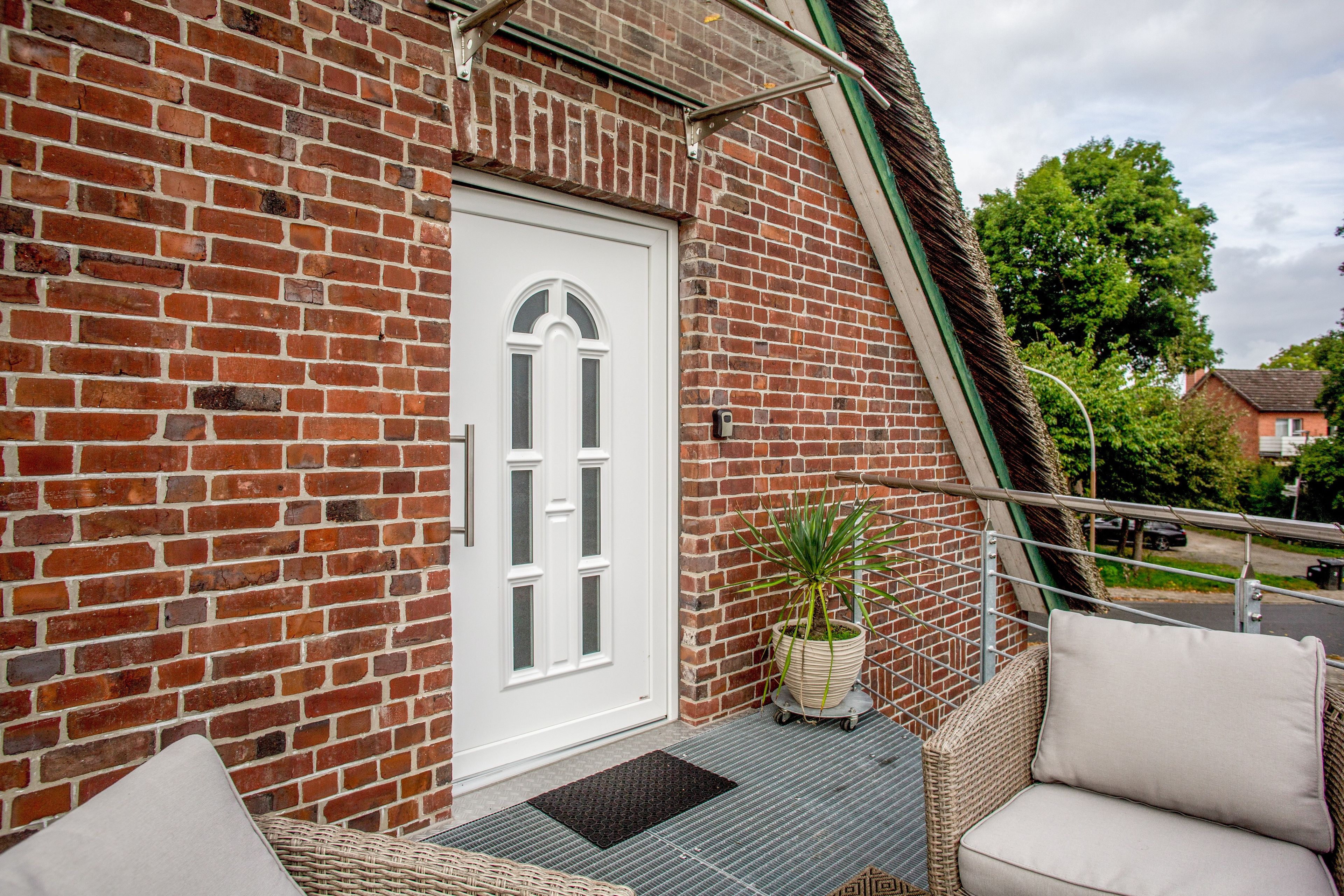 A brick building with a white door, a small balcony with wicker furniture, and a potted plant.