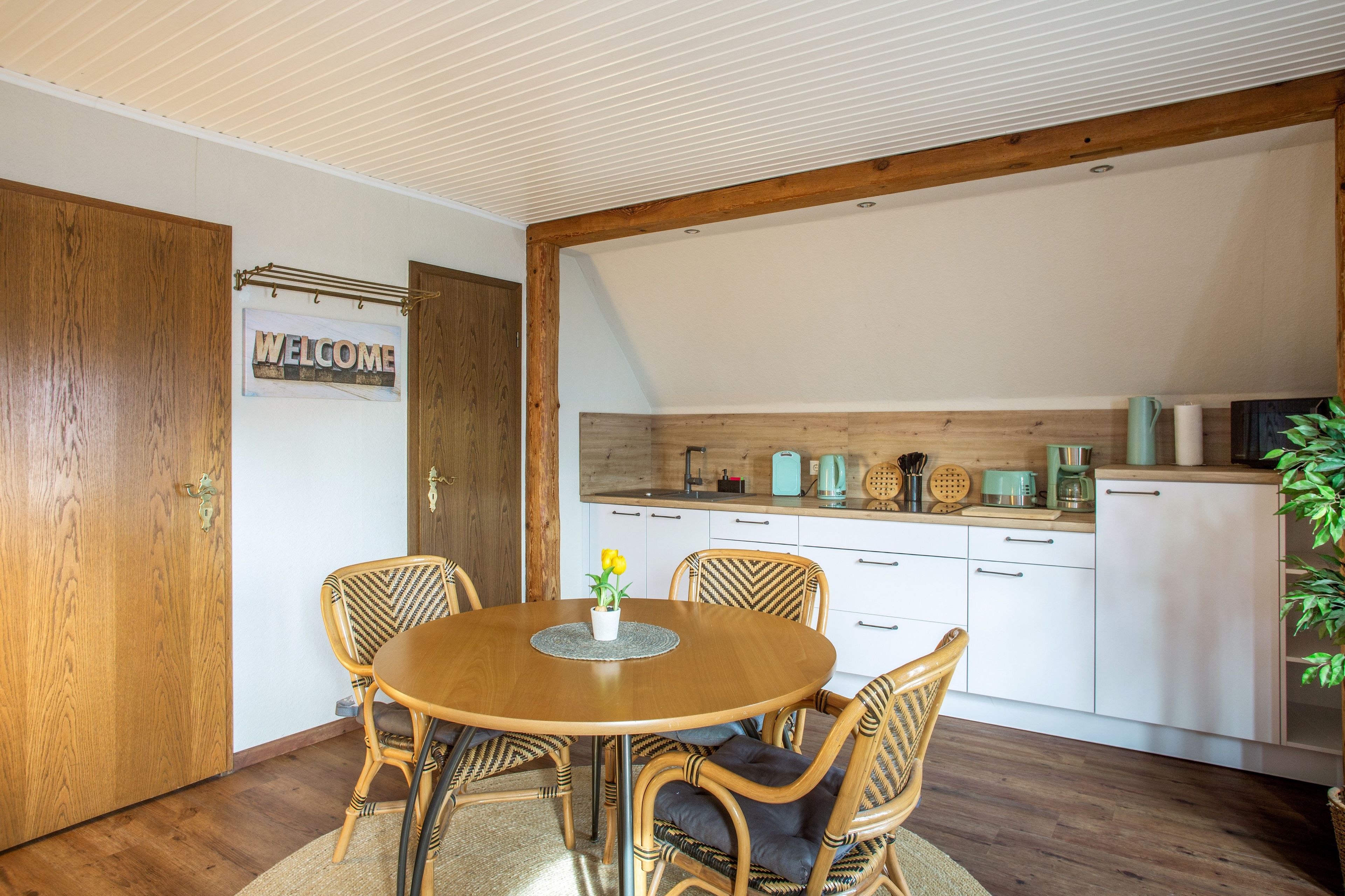 Un coin repas chaleureux avec une table et des chaises en bois, un panneau d’accueil et une cuisine dotée de meubles blancs et d’un plafond en bois.