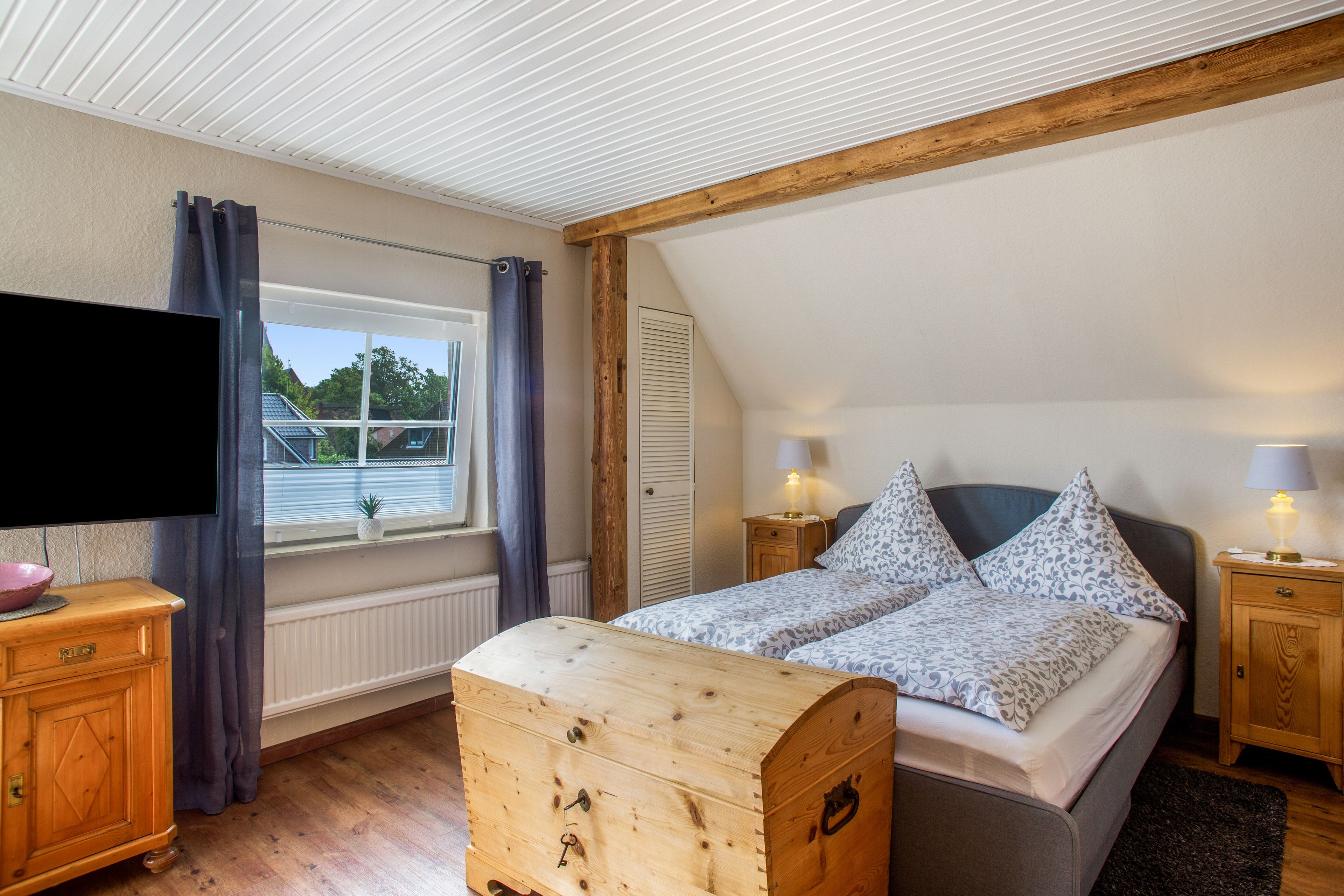 Une chambre à coucher comprenant un lit, une tête de lit en bois, des lampes de chevet, une commode, un téléviseur et une fenêtre avec des rideaux.