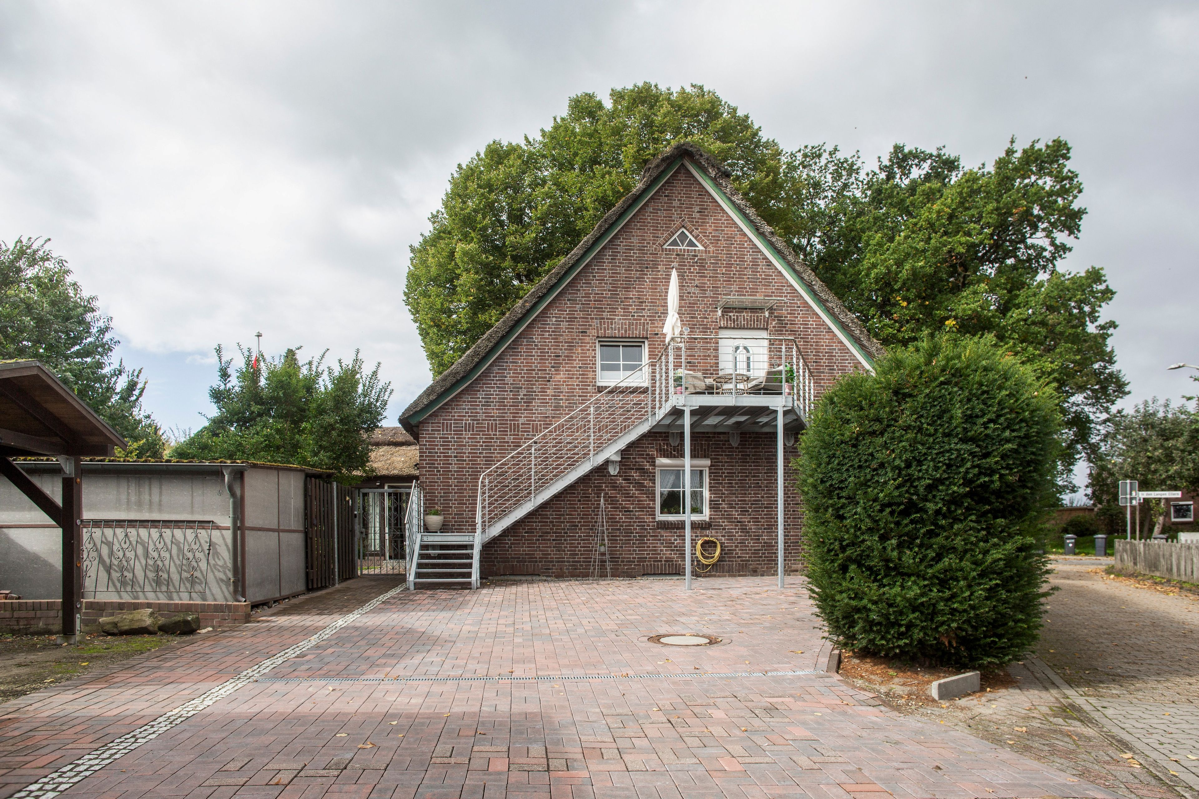 Un bâtiment en briques avec un escalier menant à un balcon, entouré d’arbres et bordé d’une allée pavée.