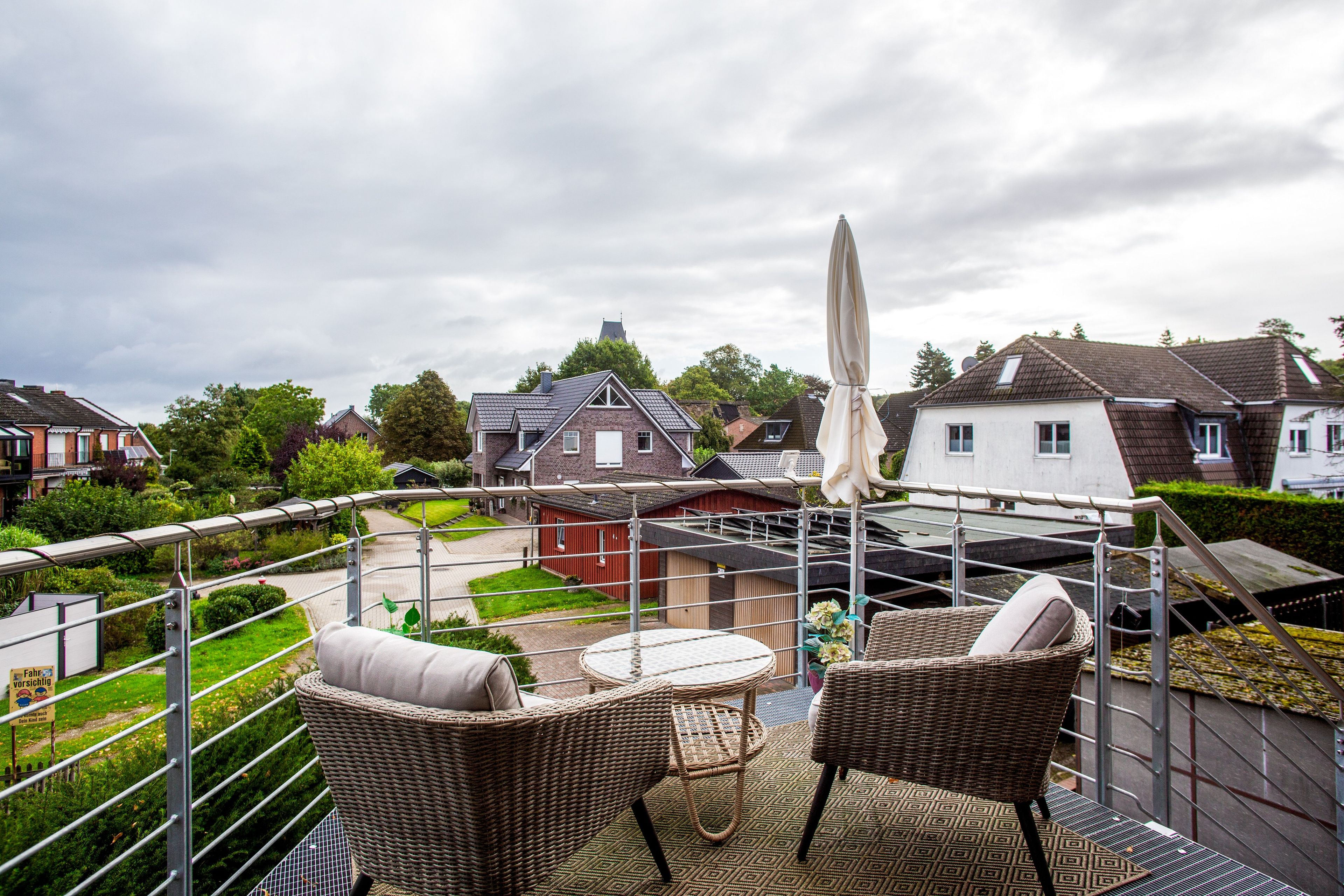Un balcon agrémenté de meubles en osier, d’un parasol blanc et offrant une vue sur des maisons et de la verdure.