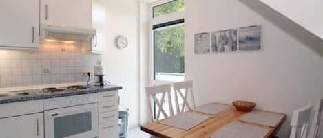 Een moderne keuken met witte kasten, een ingebouwde oven en een koffieapparaat. Er is een eettafel met stoelen en een raam met een kunstwerk erboven.