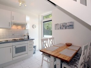 A modern kitchen with white cabinets, a built-in oven, and a coffee machine. There is a dining table with chairs and a window with artwork above it. - Waldstrasse - Holiday Apartment 26 (Wyk auf Foehr)