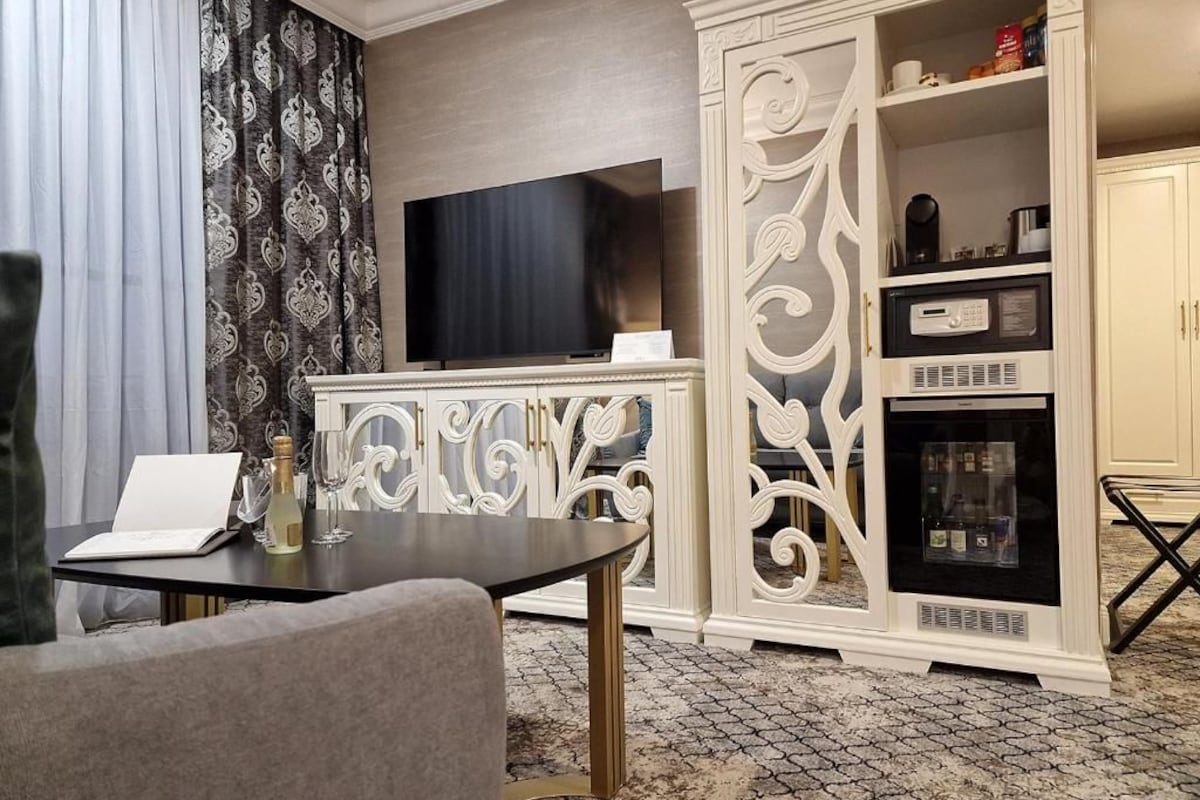 A hotel room with a flat-screen TV, a minibar, a dining area with a table and chairs, and a patterned curtain.
