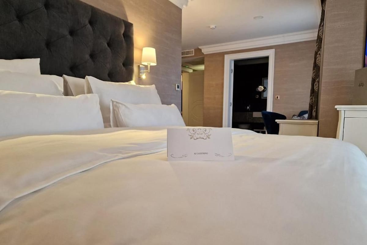A hotel room with a large bed, a headboard, a bedside table, a lamp, and a door leading to another room.