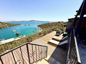 A wooden deck with a railing overlooking a marina with boats, a clear sky, and distant hills.
