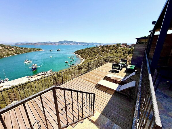 A wooden deck with a railing overlooking a marina with boats, a clear sky, and distant hills.