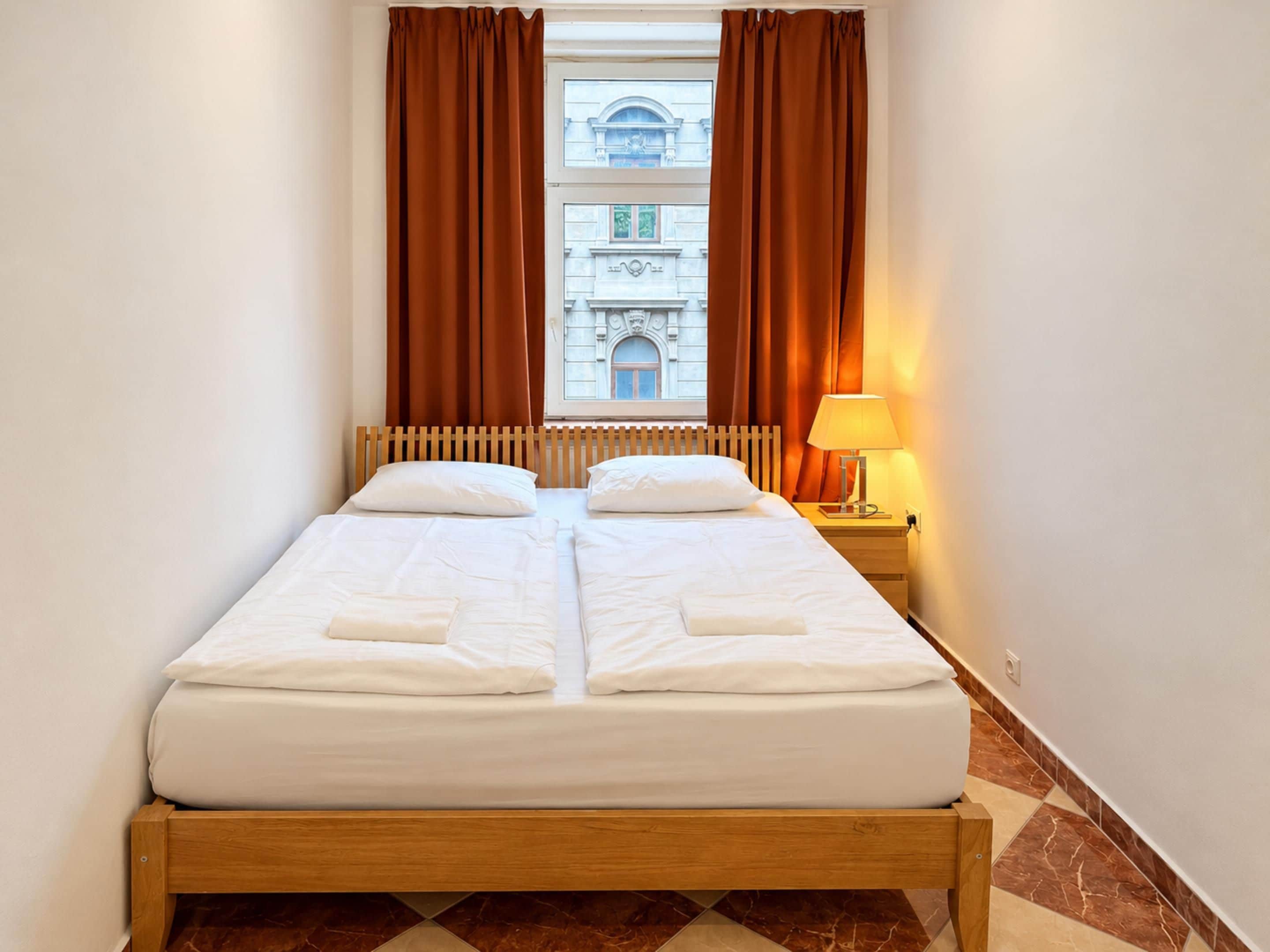 Una cama doble con sábanas blancas, cabecera de madera, mesita de noche con lámpara y ventana con cortinas marrones.