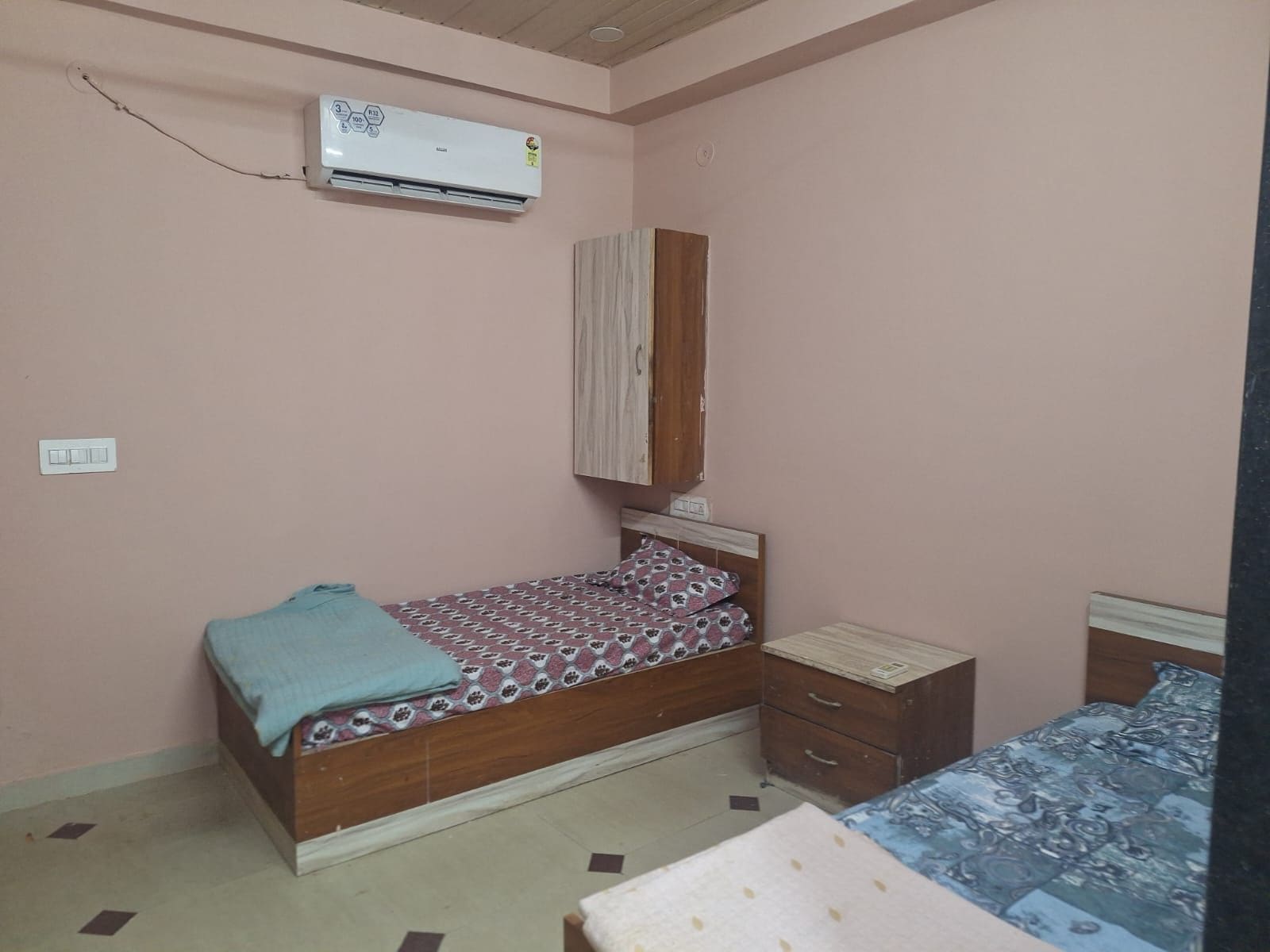 A bedroom with a single bed, a wooden wardrobe, an air conditioning unit, and a bedside table.
