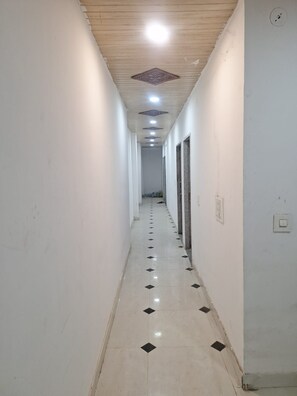 Un couloir aux murs blancs, doté d’un sol carrelé orné de motifs en losanges noirs, et d’un plafond avec des luminaires encastrés et une grille de ventilation décorative.