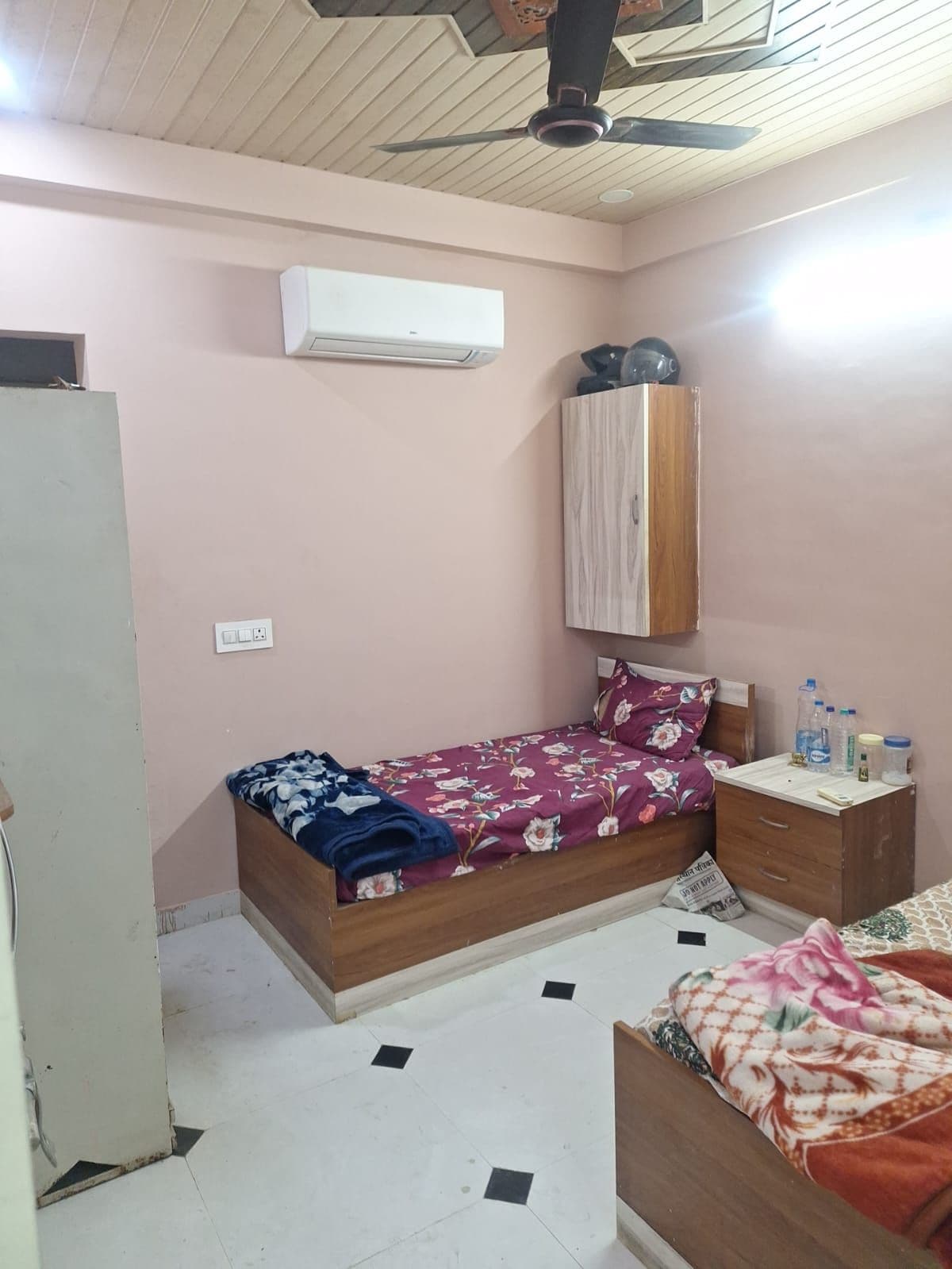 A bedroom with a single bed, a wooden wardrobe, a ceiling fan, and an air conditioning unit.