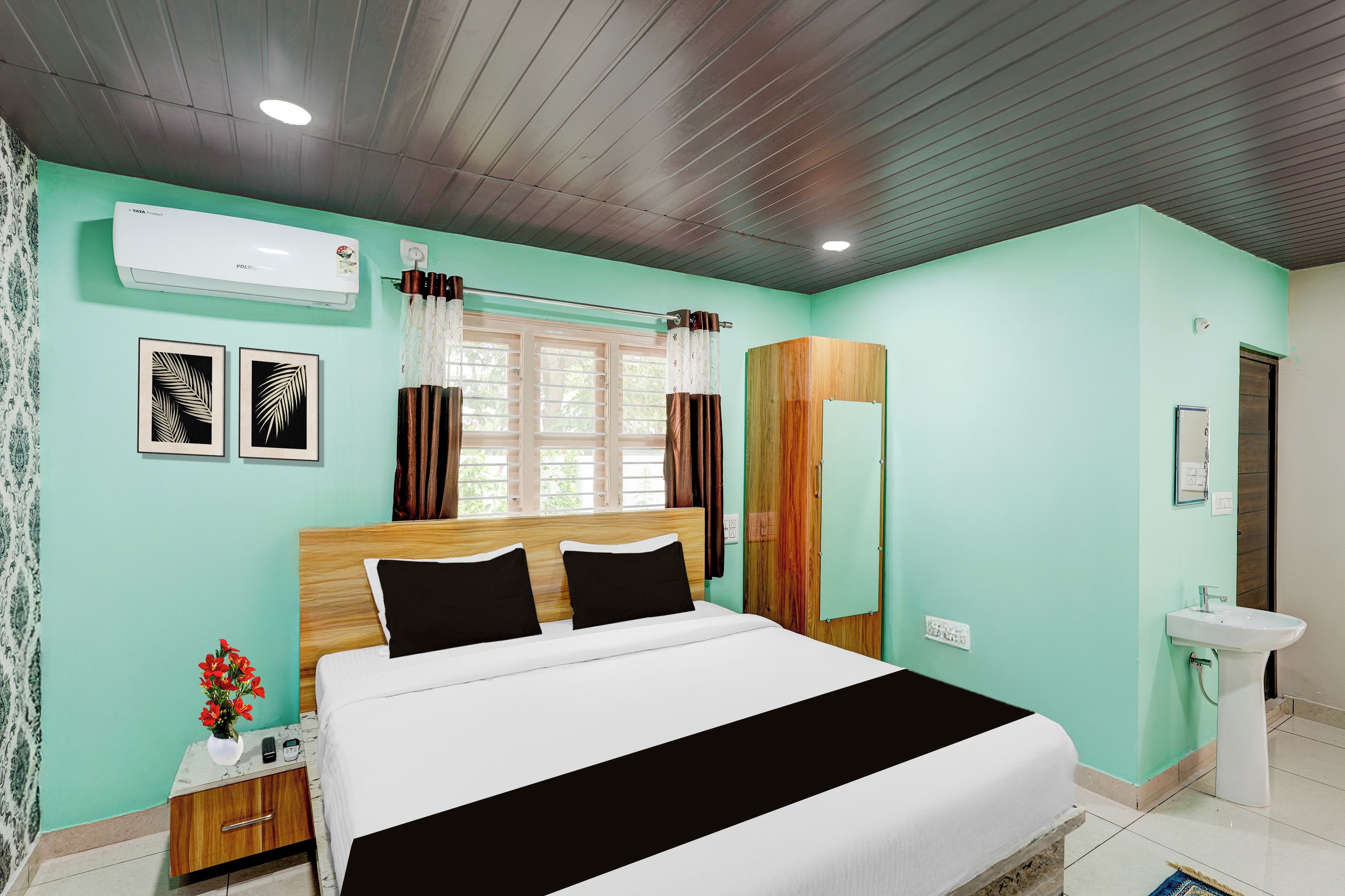 A hotel room with a bed, bedside table, a mirror, a sink, and a window with curtains.