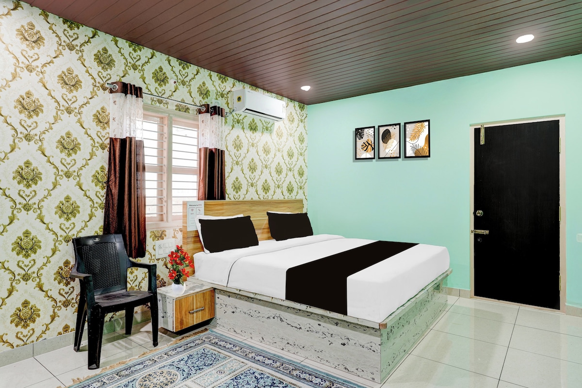 A bedroom with a bed, a chair, a bedside table with a vase of flowers, a wall-mounted air conditioner, and a door.
