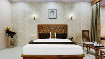 Habitación de hotel con una cama grande, cabecera de madera, mesita de noche, una silla, una mesa con hervidor y tazas, y un jarrón con flores.