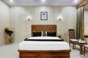 Habitación de hotel con una cama grande, cabecera de madera, mesita de noche, una silla, una mesa con hervidor y tazas, y un jarrón con flores.