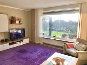 Una sala de estar con televisor, un sofá, una mesa de centro y un ventanal con vista a un jardín.