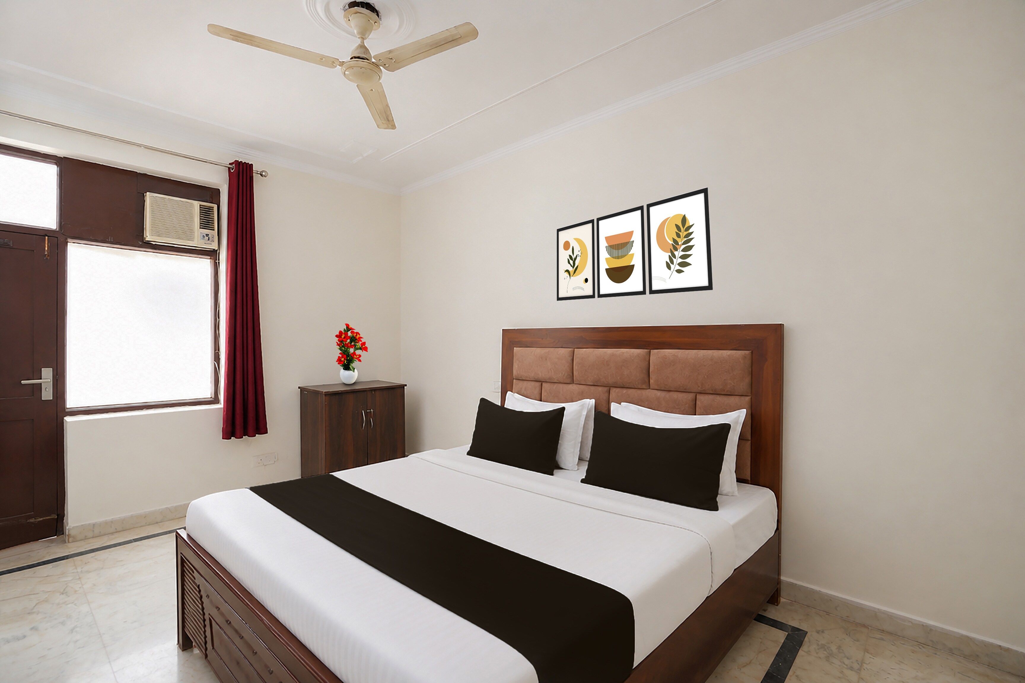 A hotel room with a bed, a ceiling fan, a window with curtains, an air conditioning unit, a small cabinet, and three framed pictures on the wall.