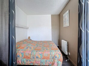 A small, single-bed room with a patterned bedspread, a radiator, and a framed picture on the wall. - Studio for 4 ppl near the sea (Agde)