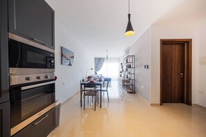 A modern kitchen with built-in appliances, a dining area with a table and chairs, and a wooden door. - Stay in this bright and modern 1-bedroom apartment in Marsaskala, Malta. Enjoy a spacious open-plan living area, fully equipped kitchen, air conditioning and a comfortable double bedroom, ideal for couples, solo travellers or longer stays. (Marsaskala)