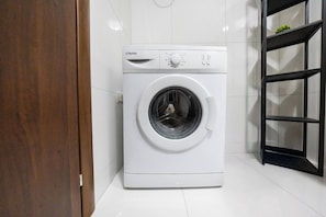 A white washing machine in a corner of a room with white tiled walls and a black shelf. - Stay in this bright and modern 1-bedroom apartment in Marsaskala, Malta. Enjoy a spacious open-plan living area, fully equipped kitchen, air conditioning and a comfortable double bedroom, ideal for couples, solo travellers or longer stays. (Marsaskala)