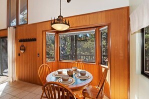A dining area with a wooden table and chairs, a chandelier, and large windows with a view of trees. - Serenity Treehouse | Forest Views | National Forest Trail & Sledding Access (Big Bear Lake)