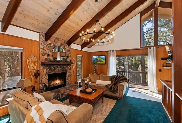 A cozy living room with a stone fireplace, wooden beams, a large window, and a comfortable seating area. - Serenity Treehouse | Forest Views | National Forest Trail & Sledding Access (Big Bear Lake)
