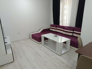 A room with a white sofa, a small white table, and a window with black curtains. - One-Bedroom Apartment-14 Filip Makedonski 49B (Plovdiv)